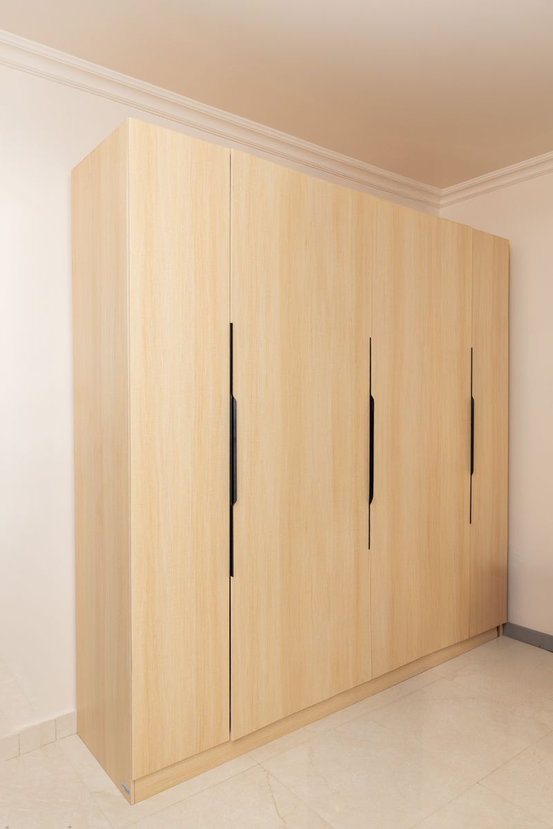 Modern light wood wardrobe with sleek black handles in a bright bedroom setting.